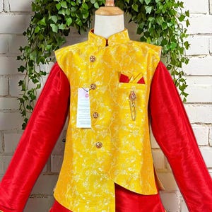 May include: A vibrant yellow and red outfit for a child. The ensemble includes a yellow vest with floral patterns and gold buttons, a red long-sleeved shirt, and red and yellow pants. A decorative pin and pocket square add detail.
