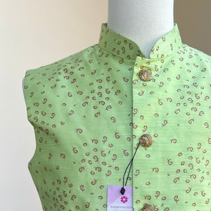 May include: A light green, sleeveless vest with a mandarin collar and gold-colored buttons. The vest features a repeating pattern of small, brown, circular shapes. A tag is attached to the vest.