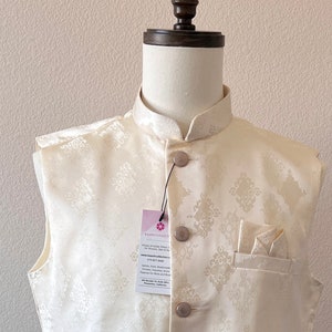 May include: A cream-colored sleeveless vest with a mandarin collar and a white patterned design. The vest has five buttons down the front and two pockets. A small white pocket square is in the top right pocket. The vest is on a white mannequin.