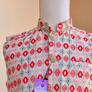 May include: A white, red, and green patterned waistcoat with gold buttons. The waistcoat is on a white mannequin. The tag on the waistcoat reads "KAASH COLLECTION House of Indian Ethnic Wear for Women, Men & Kids www.kaashcollection.com 415-827-9482 Sarees, Suits, Readymade blouses, Dupattas, Kurta Pajamas for Mens and Boys 405 Boulder Ct, Suite 200-4 Pleasanton, California."
