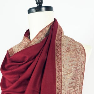 May include: A draped burgundy shawl with a detailed border. The border features a beige background with intricate patterns in a reddish-brown hue. The shawl is displayed on a mannequin, showcasing its flowing fabric and design.