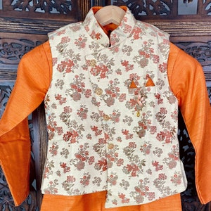 May include: A two-piece outfit for a young boy, featuring a long-sleeved orange shirt and a cream-colored vest with a floral pattern. The vest has a mandarin collar and a decorative brooch.