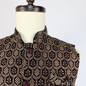 May include: A dark burgundy vest with an intricate gold floral pattern. The vest has a mandarin collar and two burgundy buttons. The fabric appears to be velvet or a similar plush material, with a subtle sheen. The vest is displayed on a mannequin.