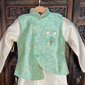 May include: A light green and gold patterned waistcoat with a white pocket square and a green and gold brooch. The waistcoat is worn over a cream colored shirt.