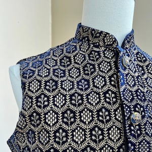 May include: A navy blue velvet waistcoat with a gold floral pattern and button closure. The waistcoat has a mandarin collar and is embroidered with a repeating pattern of flowers and leaves. The label reads "KAASH COLLECTIVE House of Indian Ethnic Wear"