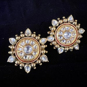 Puede incluir: Un par de pendientes de oro con un diseño circular. Los pendientes están adornados con pequeñas piedras transparentes y tienen un borde rojo alrededor del círculo central.