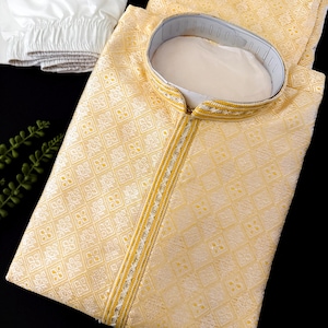May include: A light yellow kurta pajama set. The kurta features a mandarin collar and a decorative trim with gold and silver accents. The fabric has a subtle geometric pattern. White pajama pants are folded next to the kurta.