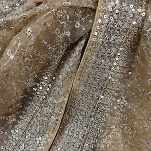 May include: Close-up of a beige fabric with intricate floral embroidery and shimmering sequins. The fabric has a delicate, sheer quality, with a detailed border along the edges. The overall effect is elegant and ornate.