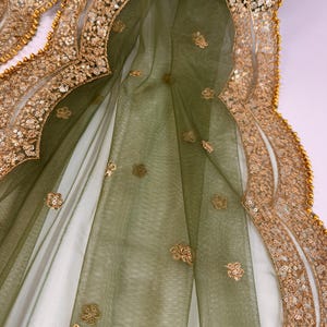 May include: Close-up of a sheer, olive green fabric with gold floral embellishments and a gold sequined border. The fabric is draped, revealing a white underlay. The gold border is detailed with small, round beads.