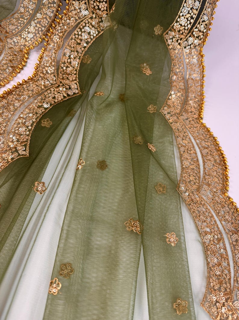 May include: Close-up of a sheer, olive green fabric with gold floral embellishments and a gold sequined border. The fabric is draped, revealing a white underlay. The gold border is detailed with small, round beads.