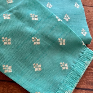 Pode incluir: Um tecido azul turquesa com um padr&atilde;o floral branco. O tecido tem uma guarni&ccedil;&atilde;o dourada ao redor das bordas.