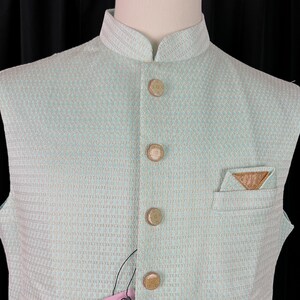 May include: A light blue sleeveless vest with a geometric pattern and brown buttons. The vest has a mandarin collar and a small pocket on the right side. The vest is made of a woven fabric and has a tag that says "KAASH COLLECTION".