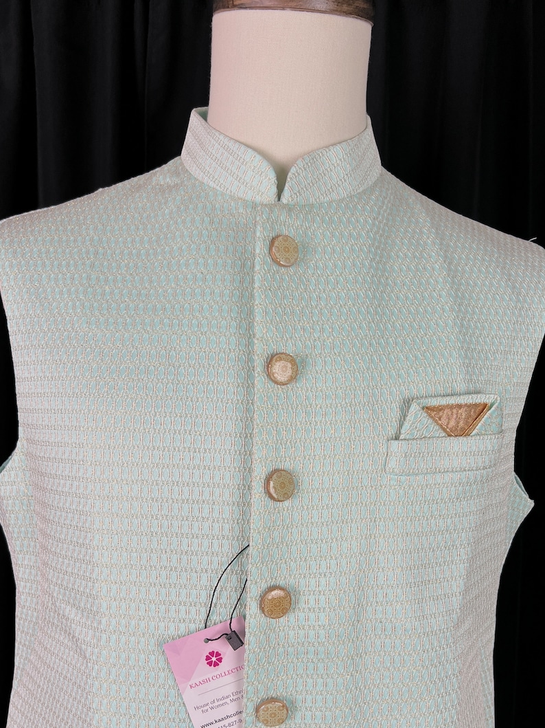 May include: A light blue sleeveless vest with a geometric pattern and brown buttons. The vest has a mandarin collar and a small pocket on the right side. The vest is made of a woven fabric and has a tag that says "KAASH COLLECTION".