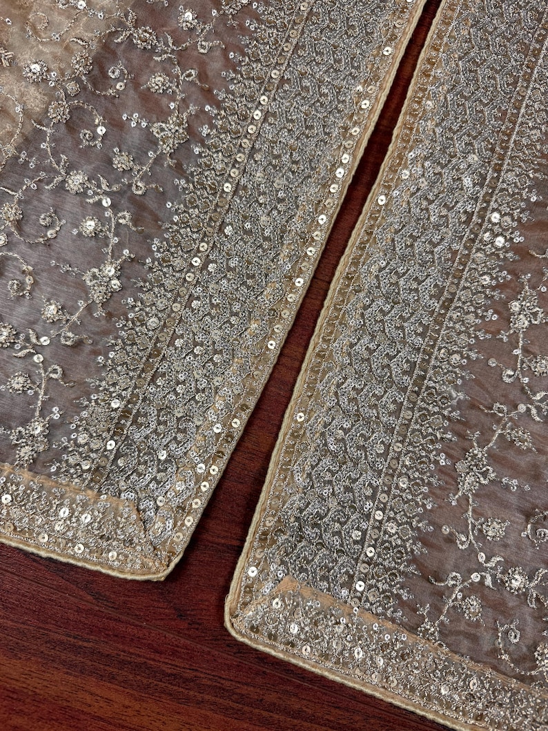 May include: Close-up of two sections of a sheer, beige fabric, heavily embellished with silver sequins and floral embroidery. The edges are trimmed with a border of sequins. The fabric is likely used for formal wear or decorative purposes.