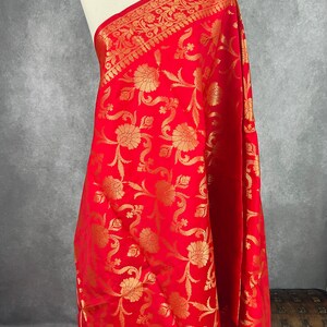 May include: A vibrant red textile with an intricate gold floral pattern. The fabric is draped over a mannequin, showcasing the detailed design and texture. The border features a decorative gold pattern.