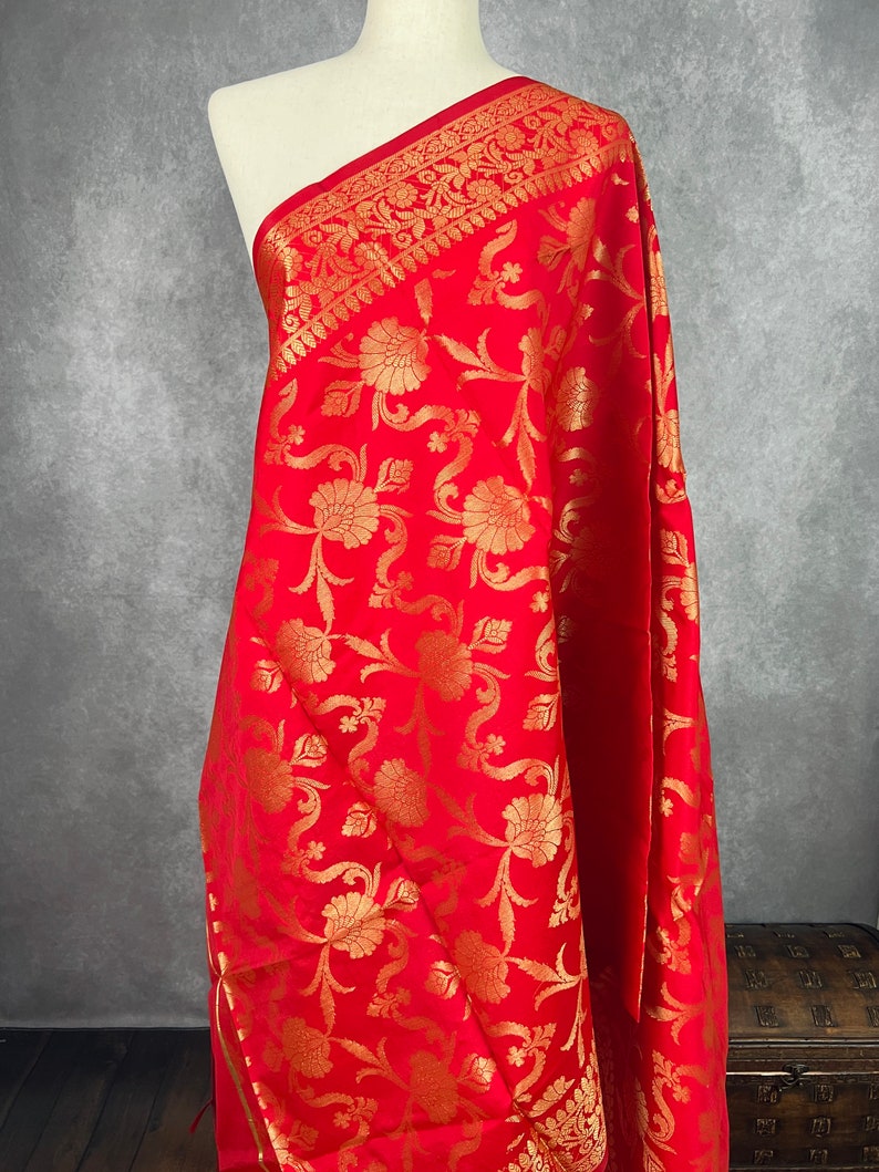 May include: A vibrant red textile with an intricate gold floral pattern. The fabric is draped over a mannequin, showcasing the detailed design and texture. The border features a decorative gold pattern.