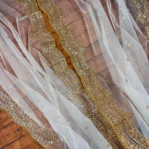 May include: White sheer fabric with a gold beaded trim. The trim features a scalloped design with a repeating pattern of small gold beads and sequins.