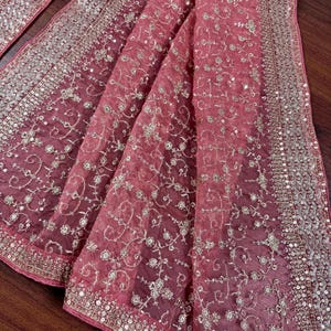 Peut inclure: Un tissu drap&eacute; et transparent dans des tons de rose et de rose, orn&eacute; de broderies et de paillettes dor&eacute;es complexes. Le tissu pr&eacute;sente un motif floral d&eacute;licat et une bordure scintillante, cr&eacute;ant une apparence luxueuse et &eacute;l&eacute;gante.