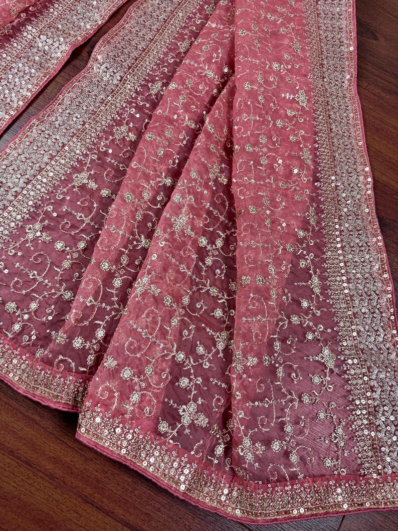 Peut inclure: Un tissu drap&eacute; et transparent dans des tons de rose et de rose, orn&eacute; de broderies et de paillettes dor&eacute;es complexes. Le tissu pr&eacute;sente un motif floral d&eacute;licat et une bordure scintillante, cr&eacute;ant une apparence luxueuse et &eacute;l&eacute;gante.