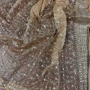 May include: A sheer, golden-toned fabric with intricate floral embroidery and shimmering sequins. The edges are adorned with a detailed border of sequins. The delicate material is draped, showcasing its flowing texture and design.