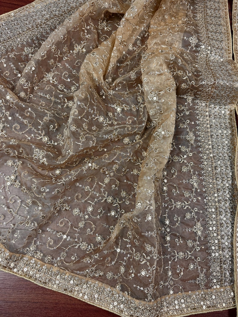 May include: A sheer, golden-toned fabric with intricate floral embroidery and shimmering sequins. The edges are adorned with a detailed border of sequins. The delicate material is draped, showcasing its flowing texture and design.