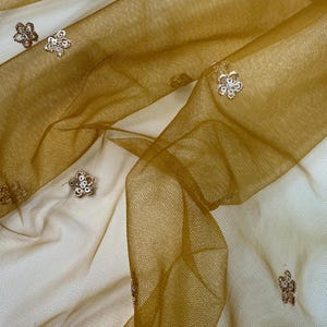 May include: Close-up of a sheer, golden-brown fabric with a delicate floral pattern. The fabric is layered, creating depth and texture. Small, embellished flowers are scattered across the surface, adding a touch of sparkle and elegance.