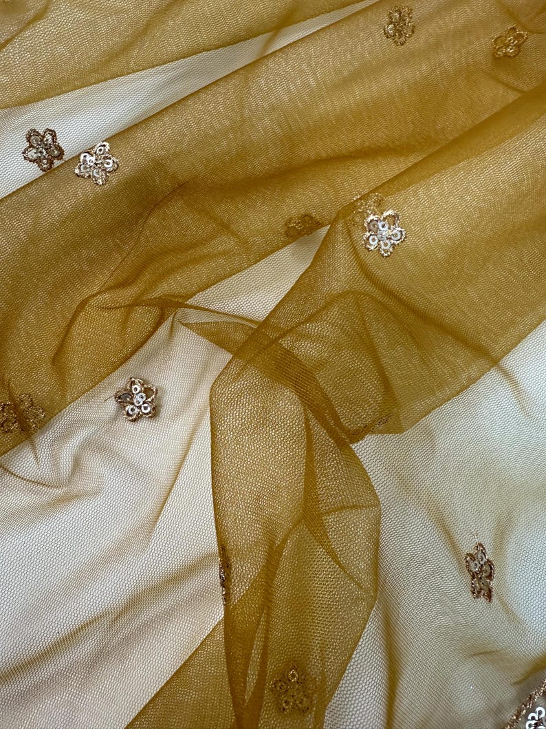 May include: Close-up of a sheer, golden-brown fabric with a delicate floral pattern. The fabric is layered, creating depth and texture. Small, embellished flowers are scattered across the surface, adding a touch of sparkle and elegance.
