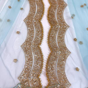 May include: A delicate light blue fabric with gold embroidery. The design includes scalloped edges and is adorned with gold sequins and small floral details. The fabric is layered over a white material, creating a layered effect.