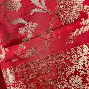 May include: Close-up of a red fabric with gold floral patterns. The fabric features a detailed design of flowers and leaves, with a decorative border along the edge. The gold patterns contrast with the red background, creating a rich visual texture.
