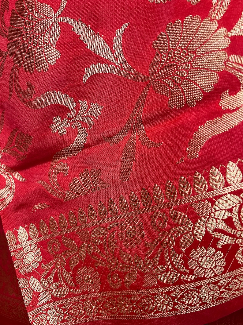 May include: Close-up of a red fabric with gold floral patterns. The fabric features a detailed design of flowers and leaves, with a decorative border along the edge. The gold patterns contrast with the red background, creating a rich visual texture.