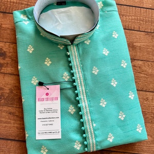 Pode incluir: Uma kurta azul turquesa com padr&otilde;es de diamantes bordados em branco e uma borda branca. A kurta tem colarinho e fecho de bot&otilde;es. O tecido &eacute; texturizado e tem um brilho sutil. A kurta est&aacute; dobrada e colocada sobre uma superf&iacute;cie de madeira. A imagem inclui uma etiqueta com o texto "KAASH COLLECTION" e informa&ccedil;&otilde;es sobre a empresa.