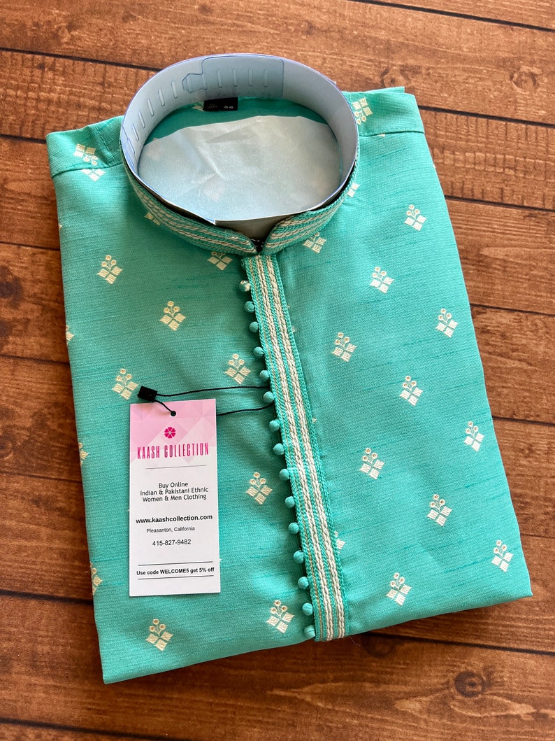 Pode incluir: Uma kurta azul turquesa com padr&otilde;es de diamantes bordados em branco e uma borda branca. A kurta tem colarinho e fecho de bot&otilde;es. O tecido &eacute; texturizado e tem um brilho sutil. A kurta est&aacute; dobrada e colocada sobre uma superf&iacute;cie de madeira. A imagem inclui uma etiqueta com o texto "KAASH COLLECTION" e informa&ccedil;&otilde;es sobre a empresa.