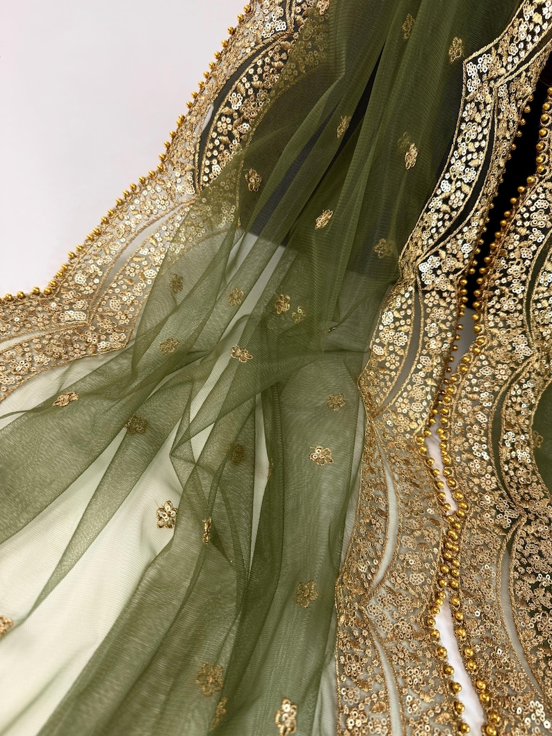 May include: Olive green sheer fabric with gold sequin and bead embellishments. The fabric has a delicate, flowing drape, with intricate gold floral patterns along the edges. The design features a combination of sequins and small beads.