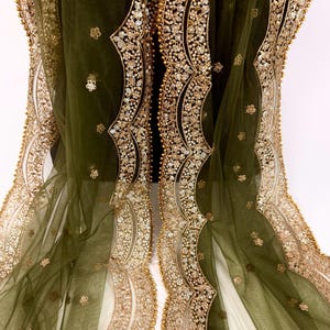 May include: Olive green sheer fabric with intricate gold sequin and bead embellishments. The edges feature a scalloped design with detailed floral patterns. The fabric is draped, showcasing the delicate texture and shimmering details.