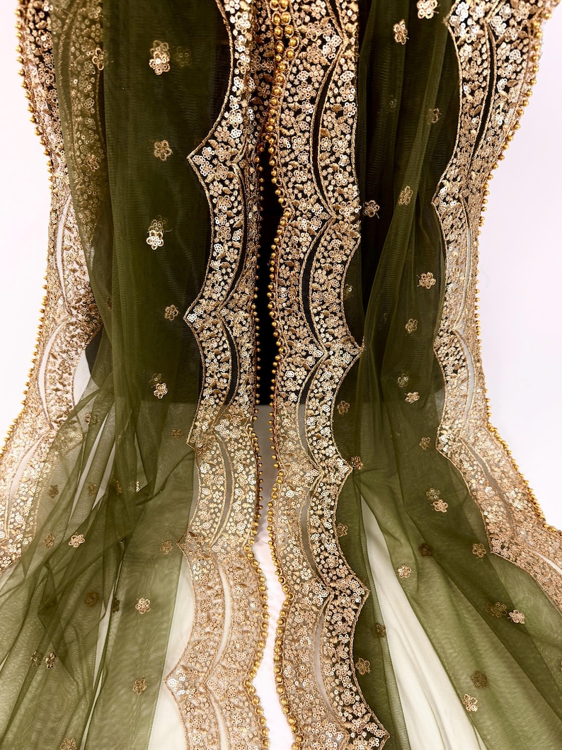 May include: Olive green sheer fabric with intricate gold sequin and bead embellishments. The edges feature a scalloped design with detailed floral patterns. The fabric is draped, showcasing the delicate texture and shimmering details.