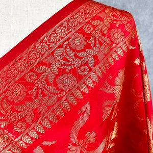 May include: Close-up of a red textile with intricate gold floral patterns. The fabric drapes over a mannequin shoulder, showcasing the detailed design. The border features a repeating pattern of gold leaves and flowers, adding to the luxurious feel.