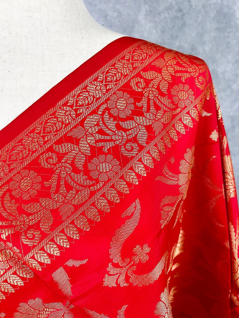 May include: Close-up of a red textile with intricate gold floral patterns. The fabric drapes over a mannequin shoulder, showcasing the detailed design. The border features a repeating pattern of gold leaves and flowers, adding to the luxurious feel.