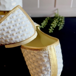 Pode incluir: Um par de sapatos baixos brancos bordados com uma borda dourada ao redor das bordas. Os sapatos s&atilde;o feitos de um tecido macio e texturizado e t&ecirc;m um ajuste confort&aacute;vel.