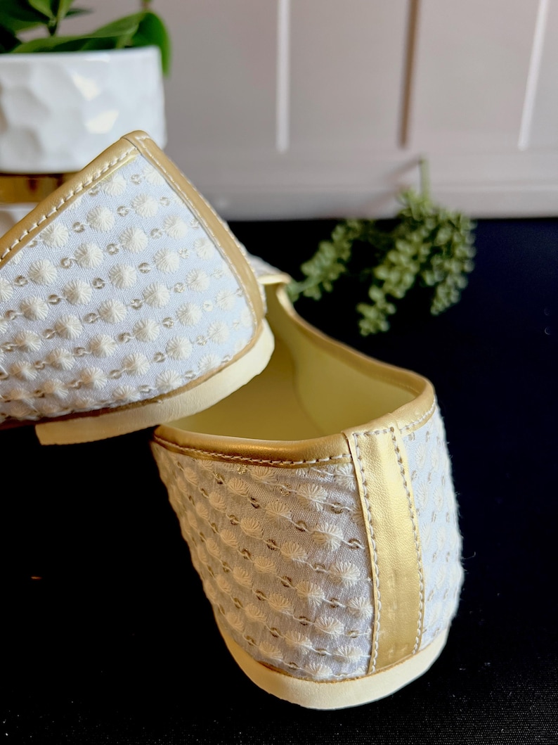 Pode incluir: Um par de sapatos baixos brancos bordados com uma borda dourada ao redor das bordas. Os sapatos s&atilde;o feitos de um tecido macio e texturizado e t&ecirc;m um ajuste confort&aacute;vel.