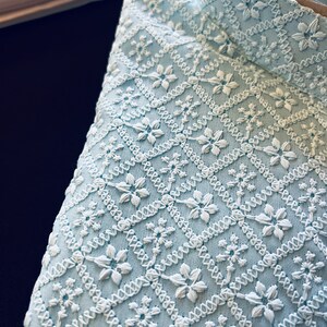 Pu&ograve; includere: Un tessuto blu chiaro con un motivo ricamato bianco di piccoli fiori e forme geometriche.