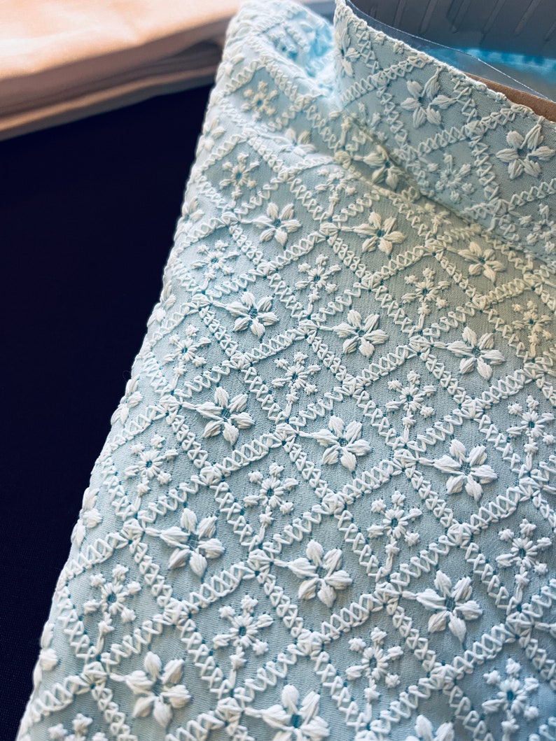 Pu&ograve; includere: Un tessuto blu chiaro con un motivo ricamato bianco di piccoli fiori e forme geometriche.