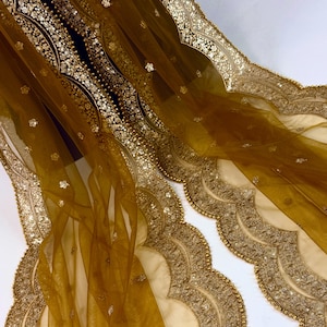 May include: A sheer, golden-yellow fabric with intricate gold embroidery and sequin detailing. The edges feature a scalloped design with beaded trim. The fabric appears to be draped, showcasing its flowing texture and ornate embellishments.