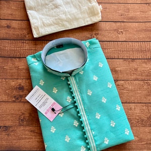 Pode incluir: Uma kurta azul turquesa com desenhos florais bordados em branco e uma borda branca com pompons brancos. A kurta tem um colar mandarim e uma etiqueta que diz "PLAIN FABRIC KURTA - DRY CLEAN ONLY - 100% COTTON - MADE IN INDIA - SIZE: M - WASH CARE INSTRUCTIONS: DRY CLEAN ONLY - DO NOT BLEACH - DO NOT TUMBLE DRY - IRON AT LOW TEMPERATURE - DO NOT WASH".