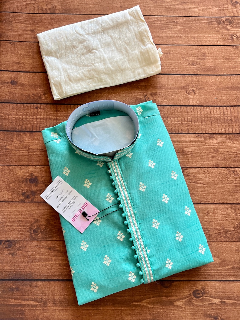Pode incluir: Uma kurta azul turquesa com desenhos florais bordados em branco e uma borda branca com pompons brancos. A kurta tem um colar mandarim e uma etiqueta que diz "PLAIN FABRIC KURTA - DRY CLEAN ONLY - 100% COTTON - MADE IN INDIA - SIZE: M - WASH CARE INSTRUCTIONS: DRY CLEAN ONLY - DO NOT BLEACH - DO NOT TUMBLE DRY - IRON AT LOW TEMPERATURE - DO NOT WASH".