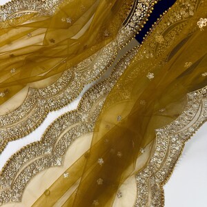 May include: Mustard yellow sheer fabric with intricate gold sequin and bead embellishments. The fabric has a scalloped edge with detailed embroidery and small floral accents. The design is rich and elegant, suitable for formal wear or special occasions.