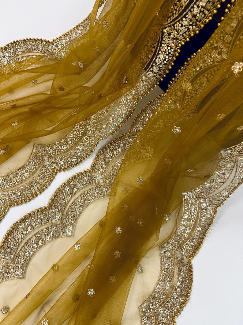 May include: Mustard yellow sheer fabric with intricate gold sequin and bead embellishments. The fabric has a scalloped edge with detailed embroidery and small floral accents. The design is rich and elegant, suitable for formal wear or special occasions.