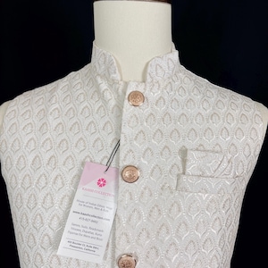 May include: White embroidered vest with a mandarin collar and gold-colored buttons. The vest features a detailed floral pattern and a small pocket. A tag with the text "KAASH COLLECTION" is attached.
