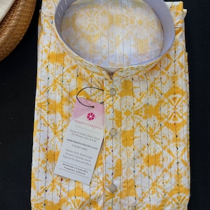 May include: A white and yellow tie-dye kurta shirt with a mandarin collar and button-down front. The shirt has a tag that reads "KAASH COLLECTION". The shirt is folded on a black surface.
