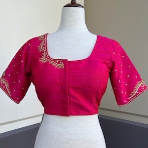 Puede incluir: Una blusa de seda rosa con bordados dorados y plateados en las mangas y el escote. La blusa tiene un escote cuadrado y mangas cortas.