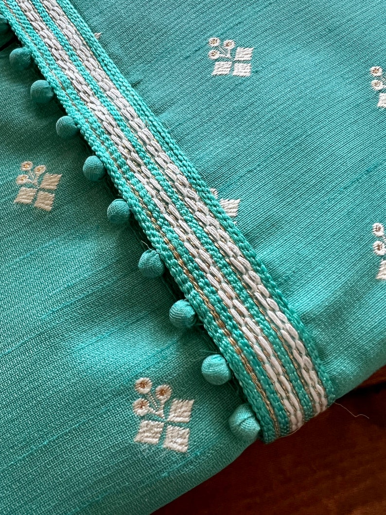 Pode incluir: Tecido turquesa com guarni&ccedil;&atilde;o branca e dourada. A guarni&ccedil;&atilde;o tem uma fila de pequenos pompons brancos ao longo da borda. O tecido tem um padr&atilde;o floral branco.